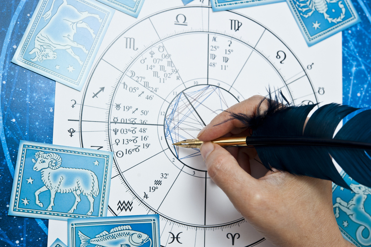Mão escrevendo com uma pena azul sobre um papel com mapa astral impresso.