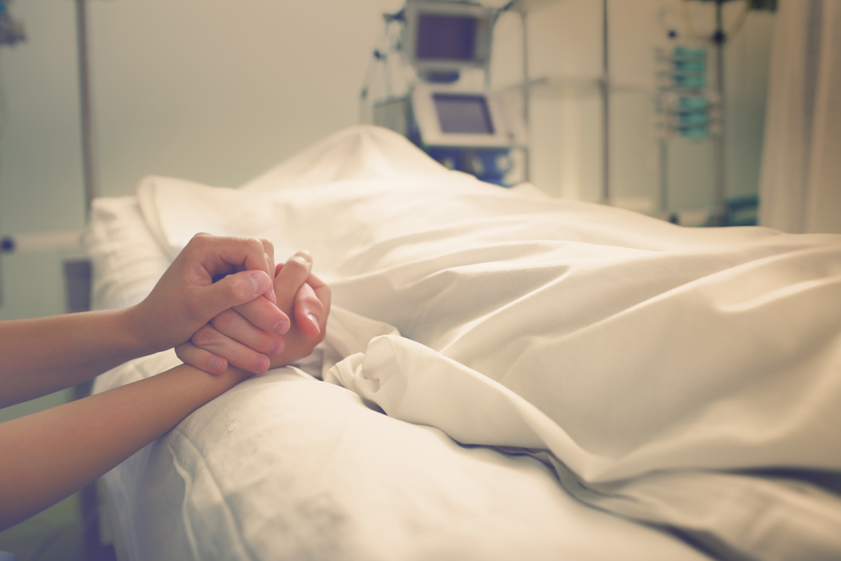 Pessoa segurando mão de enfermo em cama de hospital, representando um primo doente ou morto.