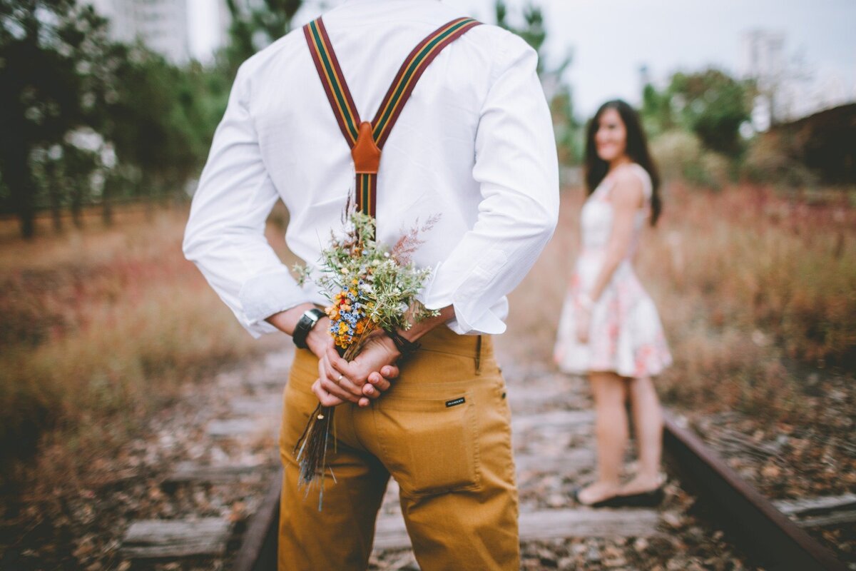Homem segurando buquê de flores escondido de mulher, que o espera, representando o amor que a Lei de Atração atrai. 