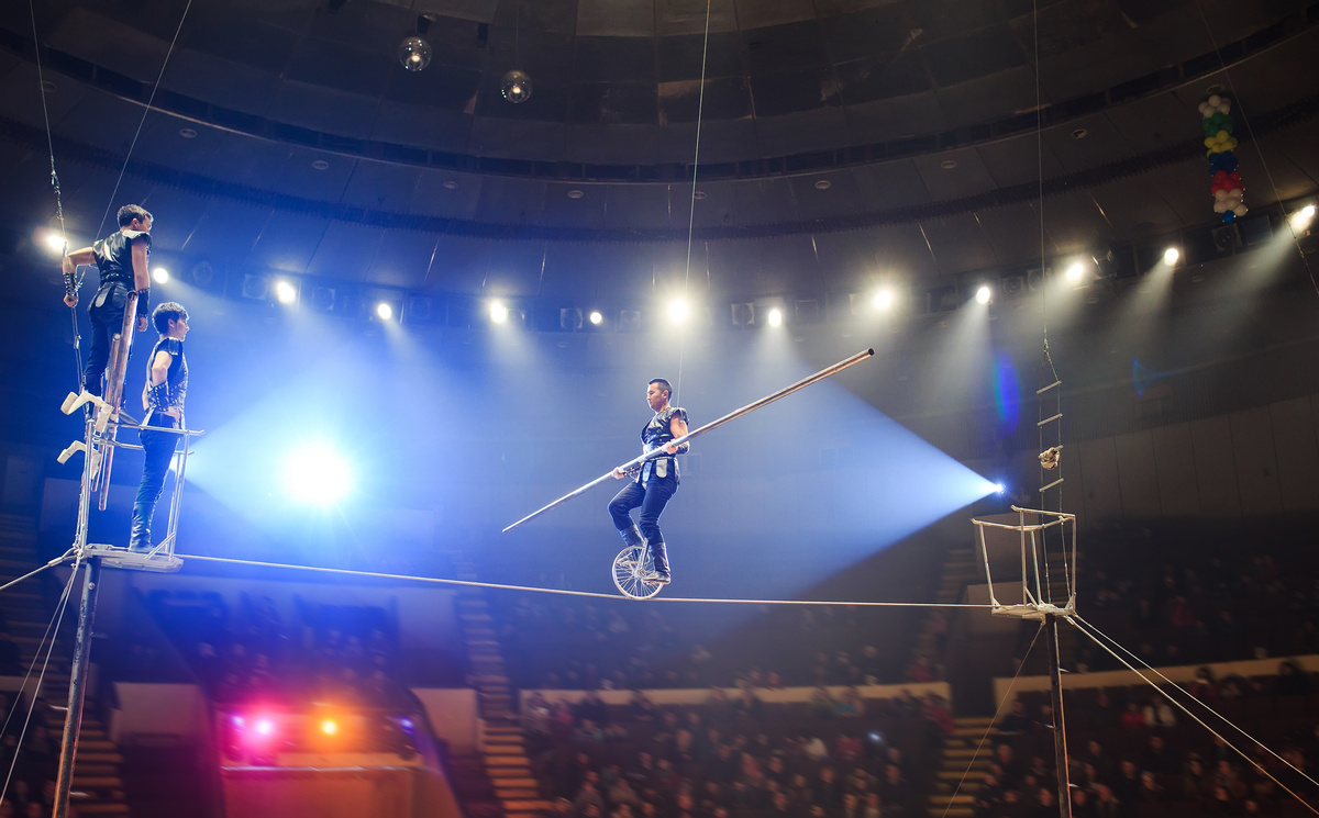 Equilibrista atravessando corda em meio a espetáculo de circo, em cenário escuro com holofotes. 