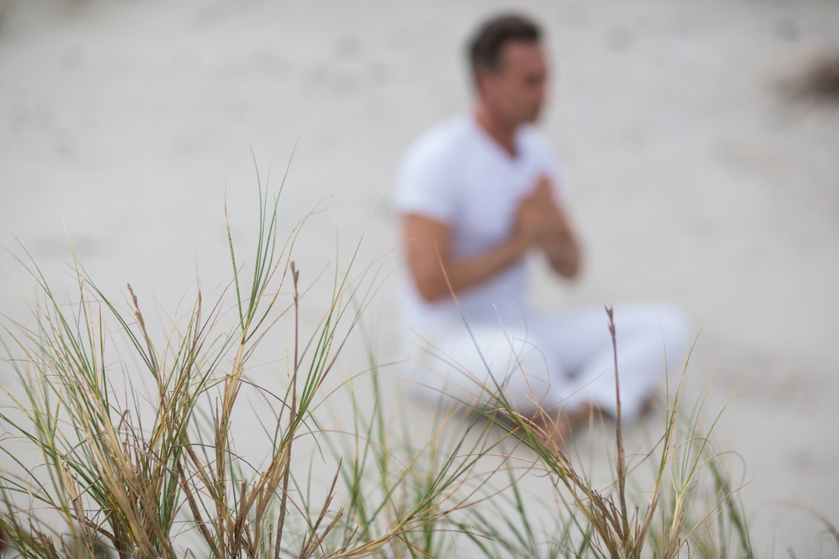 Em primeiro plano, ervas finas. Em segundo, homem vestindo branco, sentado na areia enquanto parece rezar.