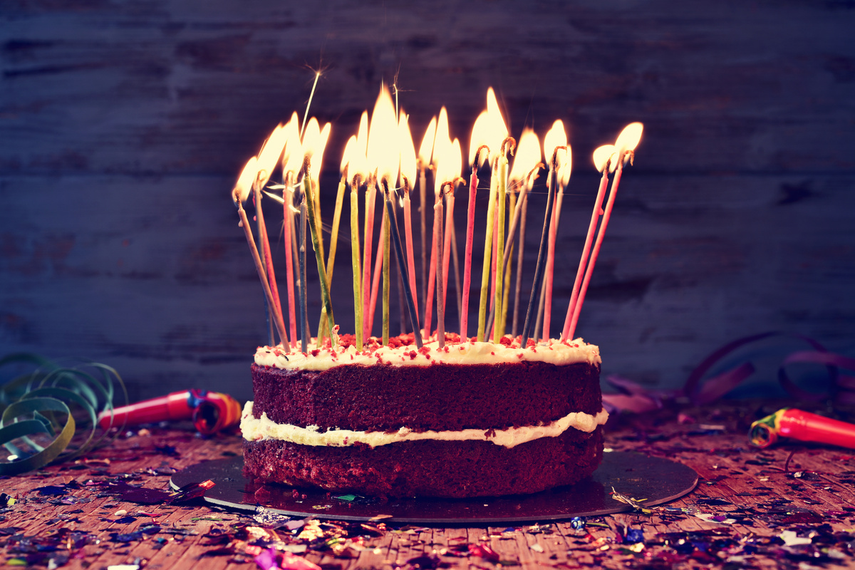 Bolo de aniversário de chocolate com muitas velas sobre mesa bagunçada.