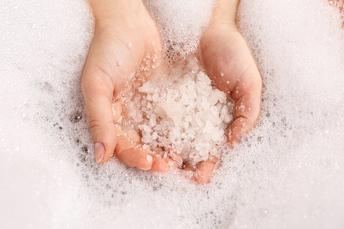 Mãos segurando punhado de sal grosso em frente à água de banho.