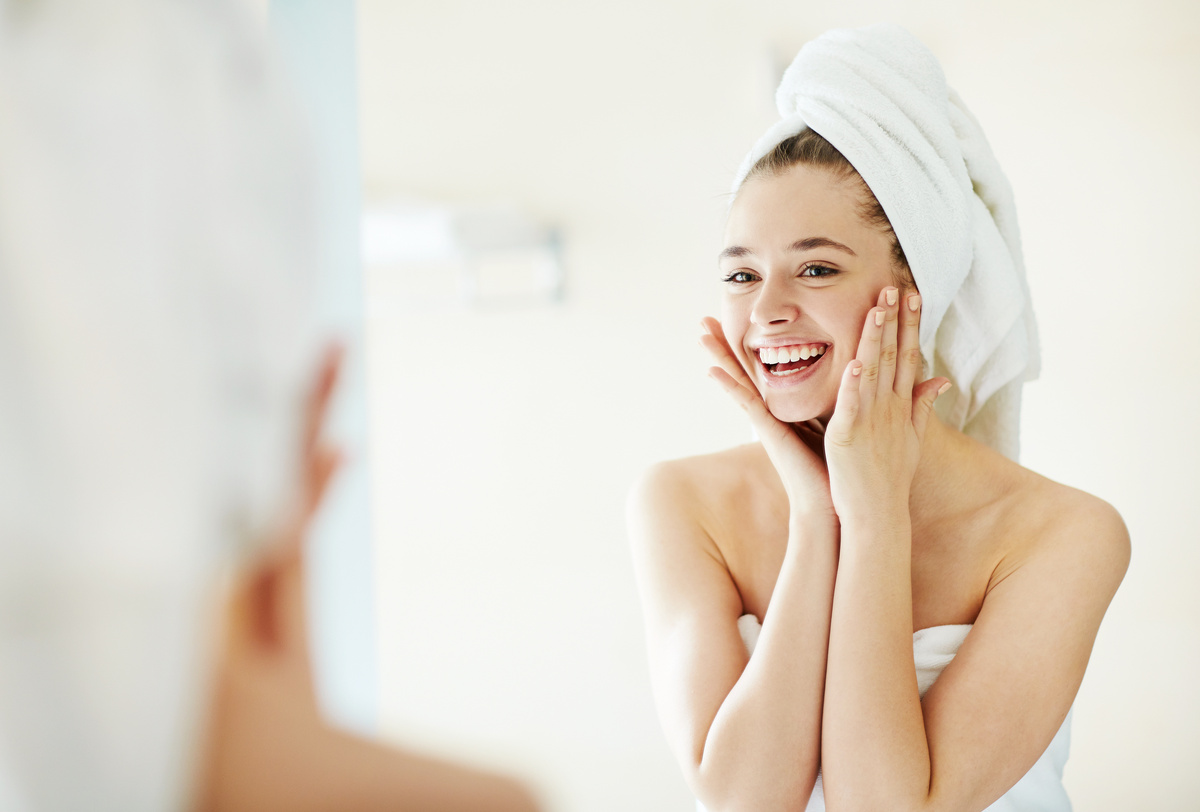 Mulher com toalha na cabeça sorrindo para o seu reflexo no espelho, simbolizando o momento após o banho para dar ânimo.