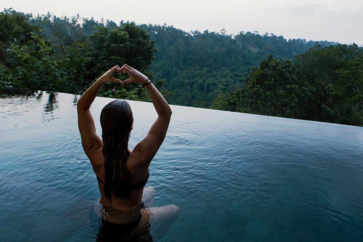 Mulher de costas, fazendo um coração com as duas mãos enquanto se senta em beirada de piscina com borda infinita.