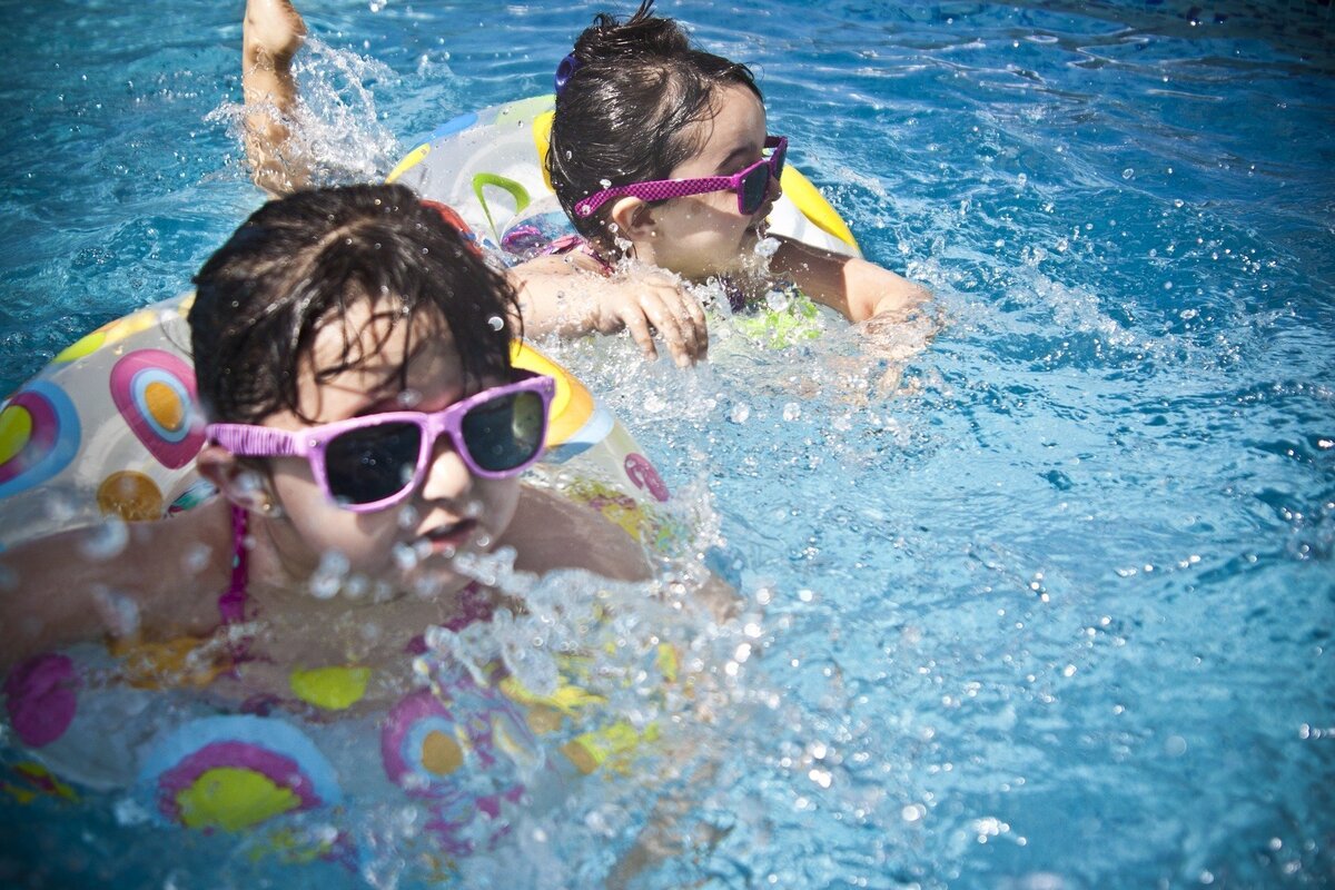 Duas crianças em boias amarelas, ambas usando óculos de sol cor-de-rosa enquanto nadam em piscina.