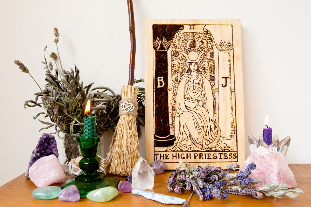 Carta A Sacerdotisa do Tarot sobre mesa de madeira com pétalas e cristais em volta.