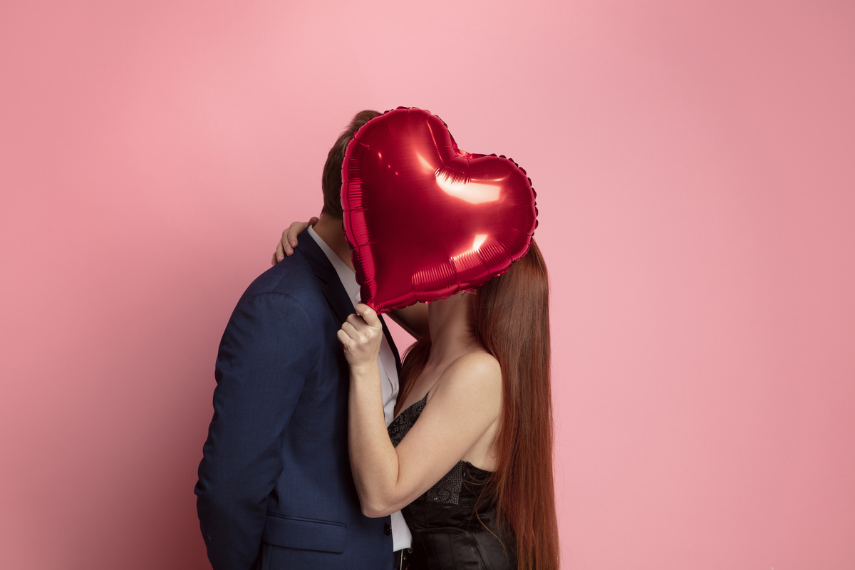 Um homem e uma mulher se beijando enquanto escondem rostos atrás de balão vermelho em formato de coração, representando o amor passional de Escorpião.
