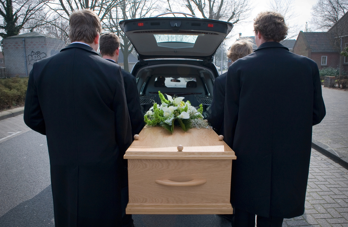 Homens carregando caixão para dentro de carro de funerária, simbolizando o falecido em um sonho.
