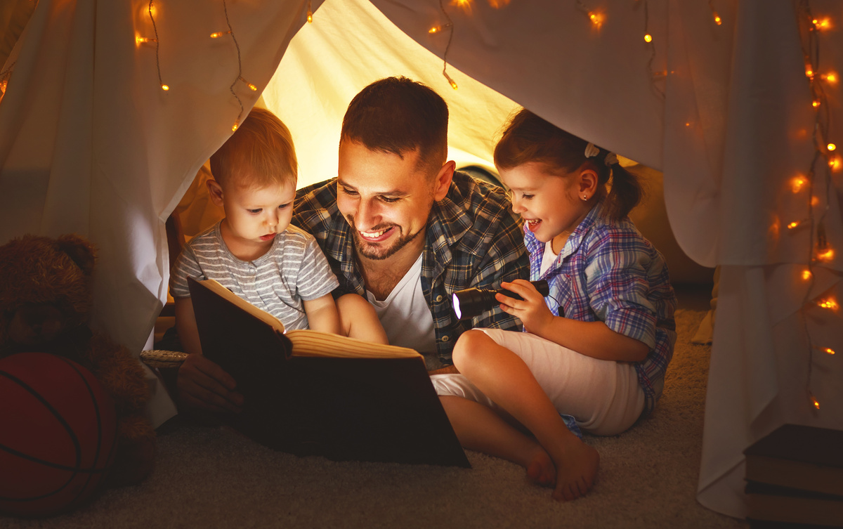 Homem em meio a duas crianças, enquanto se deitam em barraca feita com lençóis e ele lê uma história a elas, representando a dedicação do homem de Libra com a família.