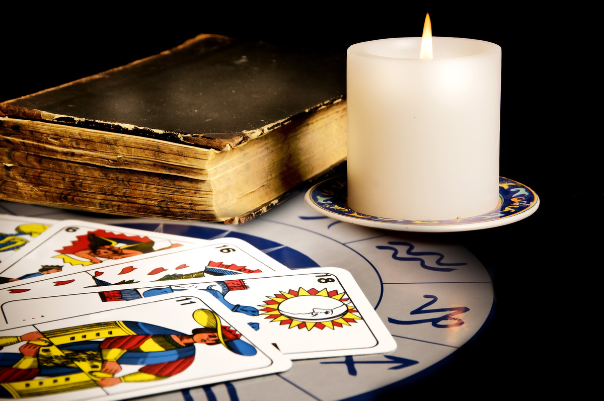 Cartas de tarot em cima de mandala astrológica em branco, enquanto ao lado de vela acesa e de um livro antigo.