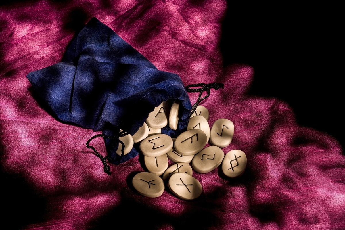 Pedras com runas gravadas saindo de dentro de pequena bolsa roxa. 