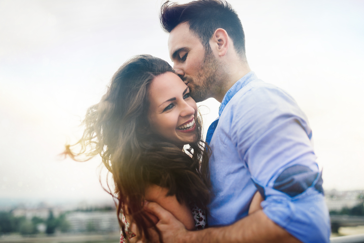 Homem beijando rosto de mulher enquanto ela sorri e o vento bate nos cabelos dela, representando o relacionamento de Gêmeos e Peixes.