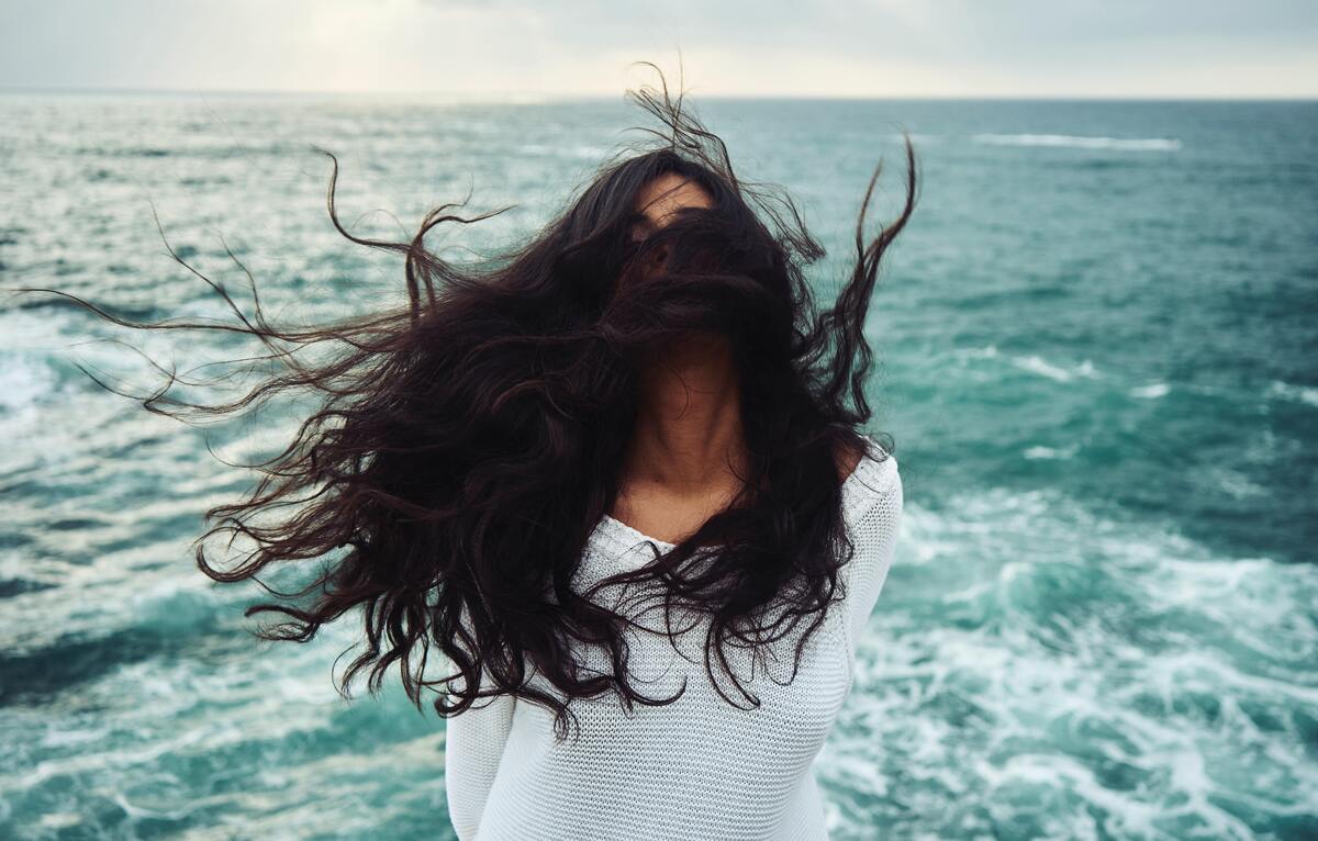 Mulher em frente ao mar com cabelo longo e escuro voando.