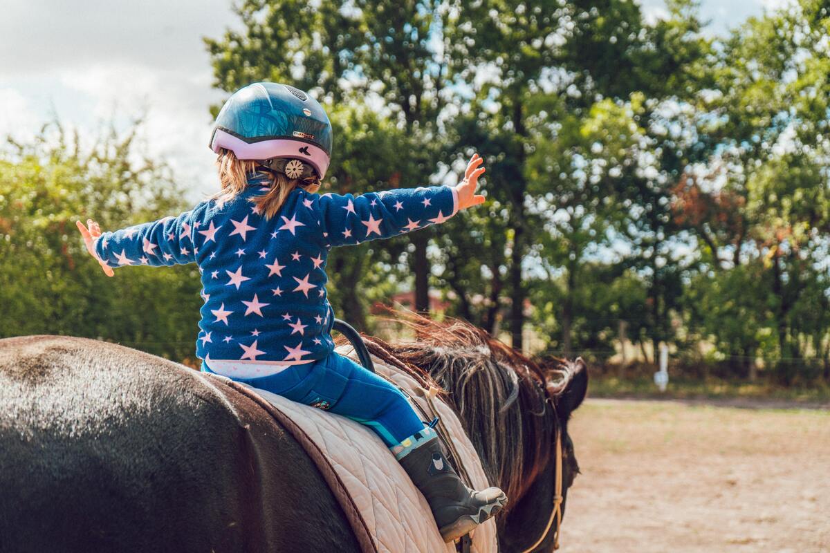 Criança em cima de um cavalo com os braços abertos e usando capacete para proteção.