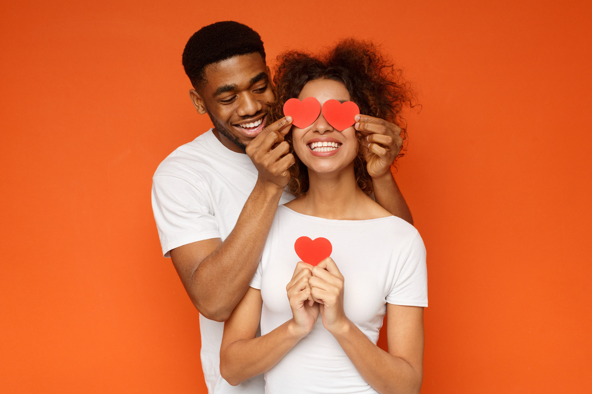 Homem por trás de mulher, colocando dois corações vermelhos em cima dos olhos dela enquanto ambos sorriem, representando o romance do taurino.