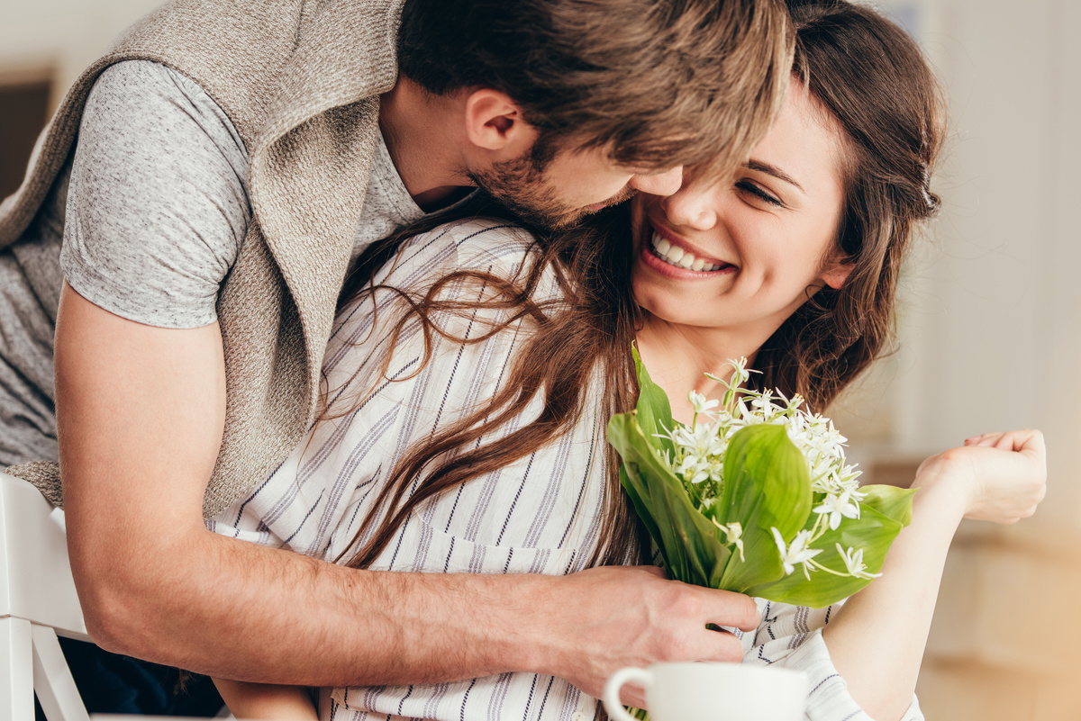Homem surpreendendo mulher por trás, abraçando-a enquanto entrega pequeno buquê de flores brancas, representando o amor do homem taurino.