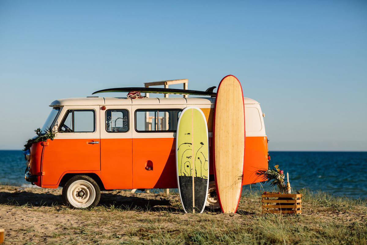 Kombi laranja estacionada na beira da praia, com pranchas de surfe apoiadas em seu corpo.