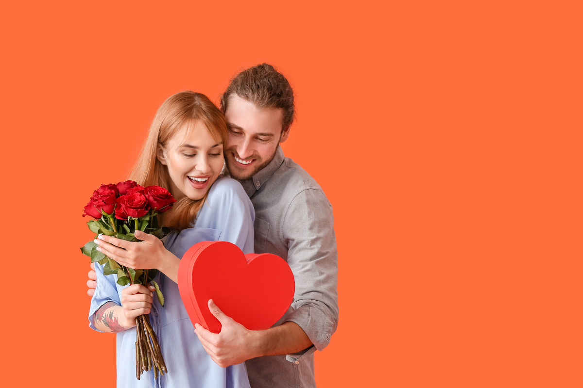 Mulher segurando buquê de flores enquanto homem a abraça por trás e entrega caixa em formato de coração, simbolizando o lado um pouco materialista de Libra no amor.