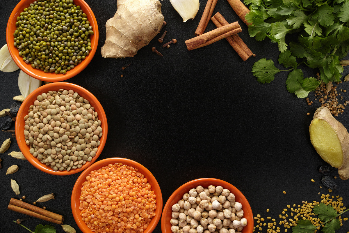 Grãos e temperos reunidos em recipientes redondos, ao lado de ervas e legumes, todos podendo ser usados na dieta da Ayurveda.