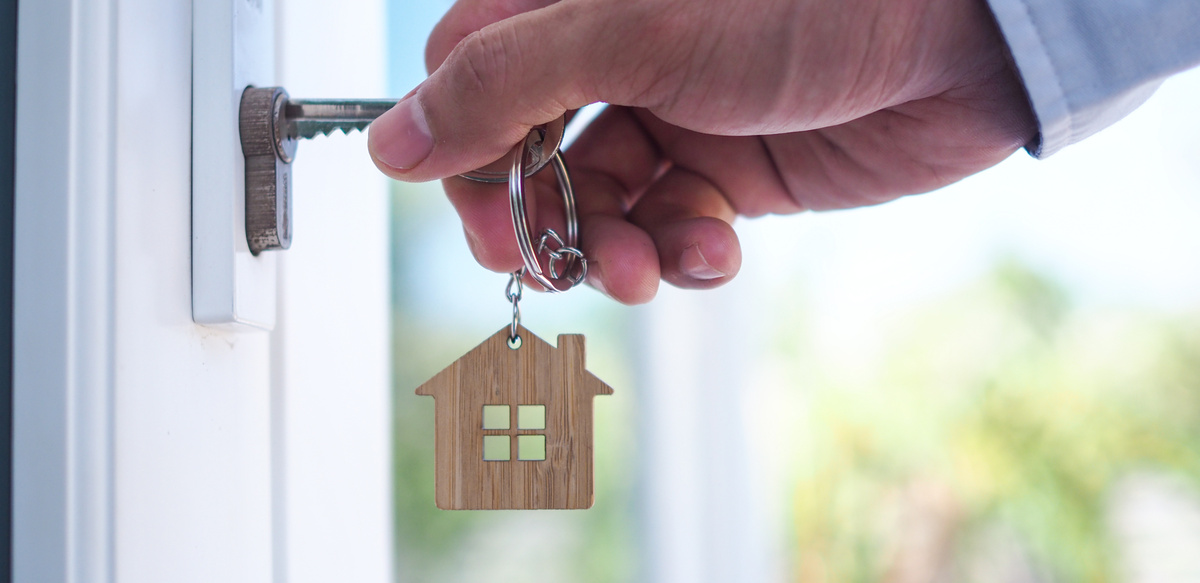 Pessoa utilizando chave, a qual possui um chaveiro de madeira de uma pequena casa, para abrir a porta.
