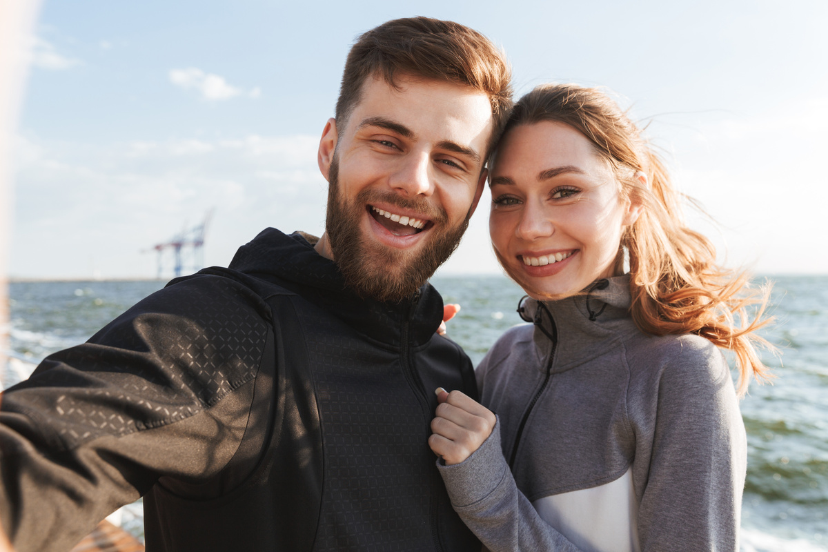 Homem e mulher posando juntos para foto que ele tira, ambos usando moletom e em frente à praia, representando o homem e a mulher de Peixes.