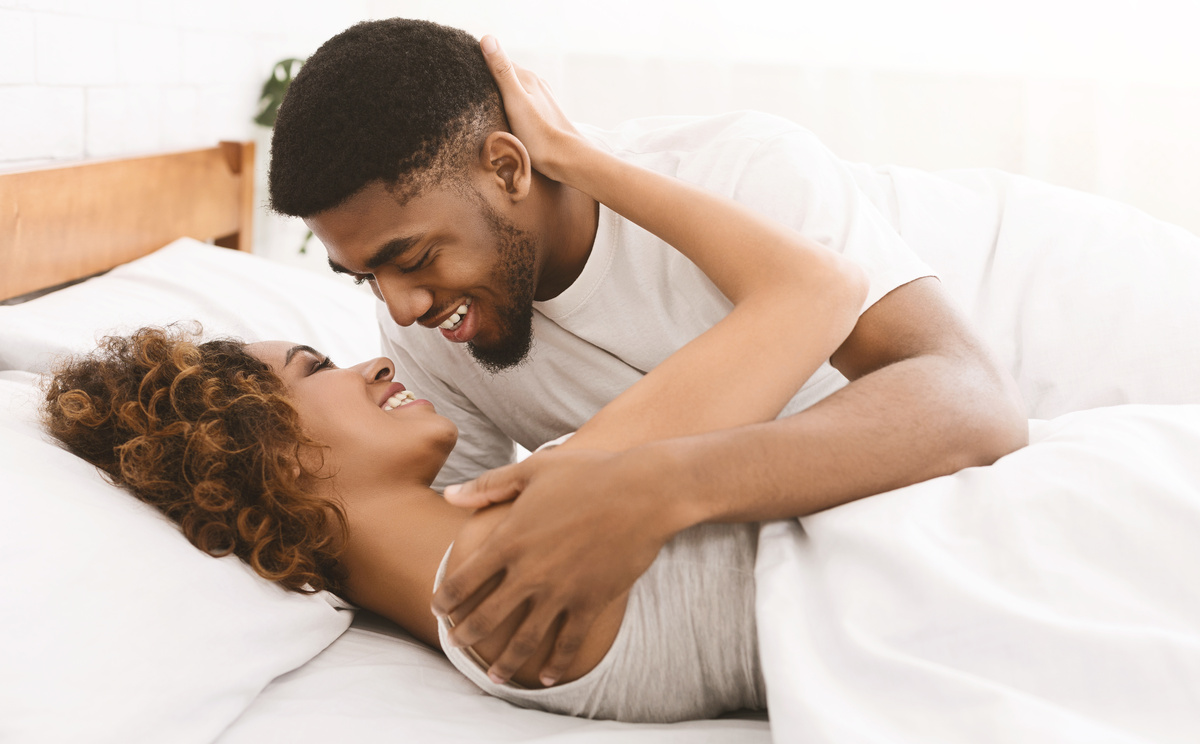 Homem e mulher deitados em cama de lençóis brancos, próximos a se beijarem, representando o comportamento do signo de Peixes durante o sexo.