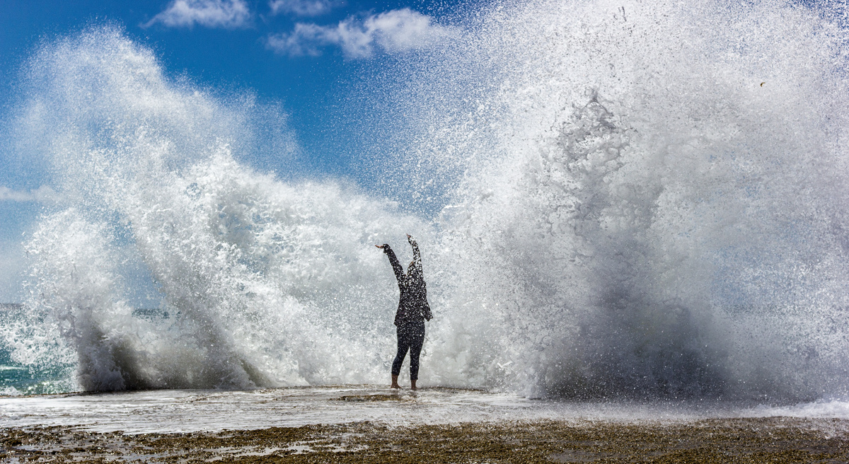 Pessoa em pé, na beira da praia, com os braços abertos, prestes a ser atingida por ondas grandes.