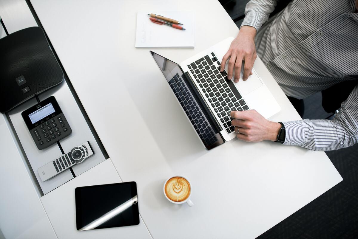 Homem usando mesa de trabalho com notebook, xícara de café, bloco de notas e celular.
