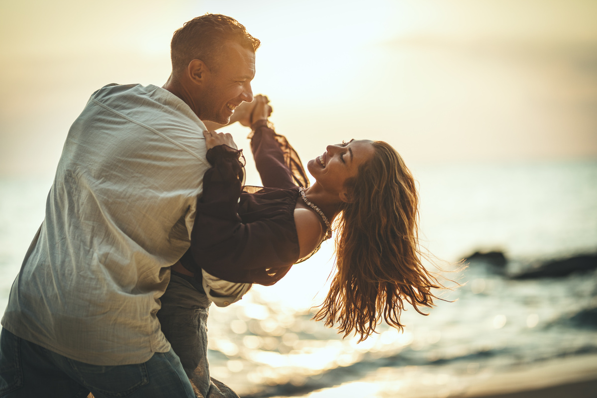 Em cenário de por do sol em frente ao mar, homem carrega mulher no colo enquanto ambos sorriem, representando o homem de Gêmeos apaixonado.