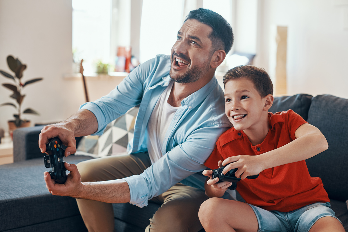 Homem e garoto jogando videogame juntos, ambos sorrindo e sentados no sofá, representando o pai e o filho de Gêmeos.