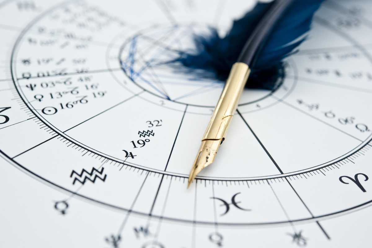 Roda do mapa astral, com desenhos das casas astrológicas, impressa em papel. Em cima dela, uma caneta preta com penas. 