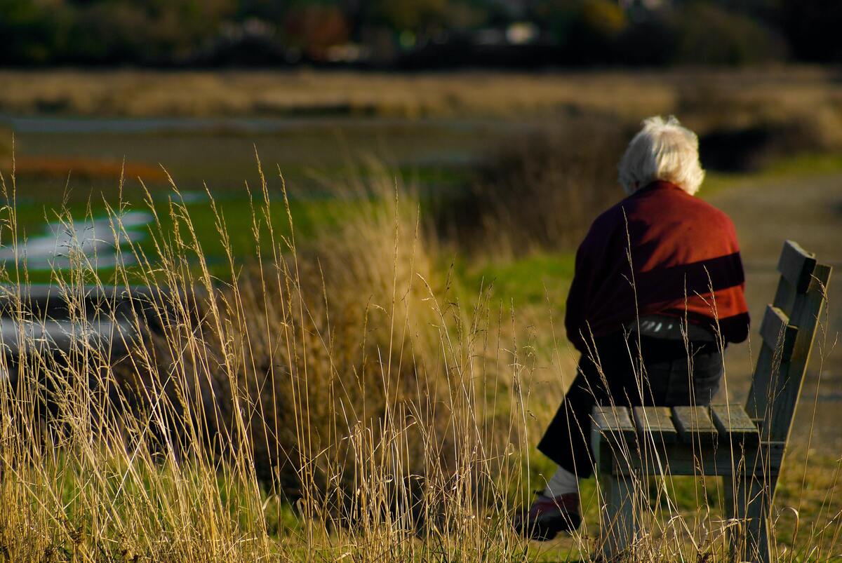Mulher idosa sentada de costas em um banco.
