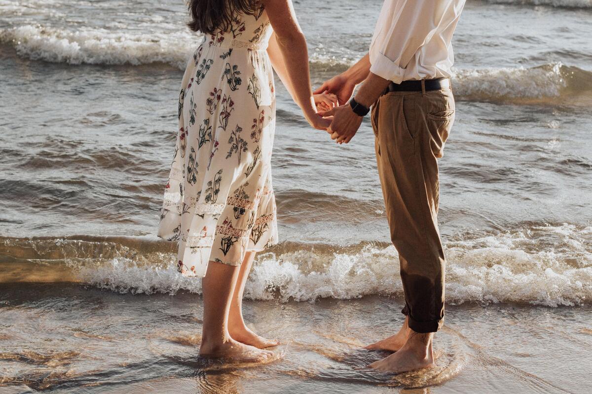 Casal em pé na praia, perto da água, de mãos dadas.