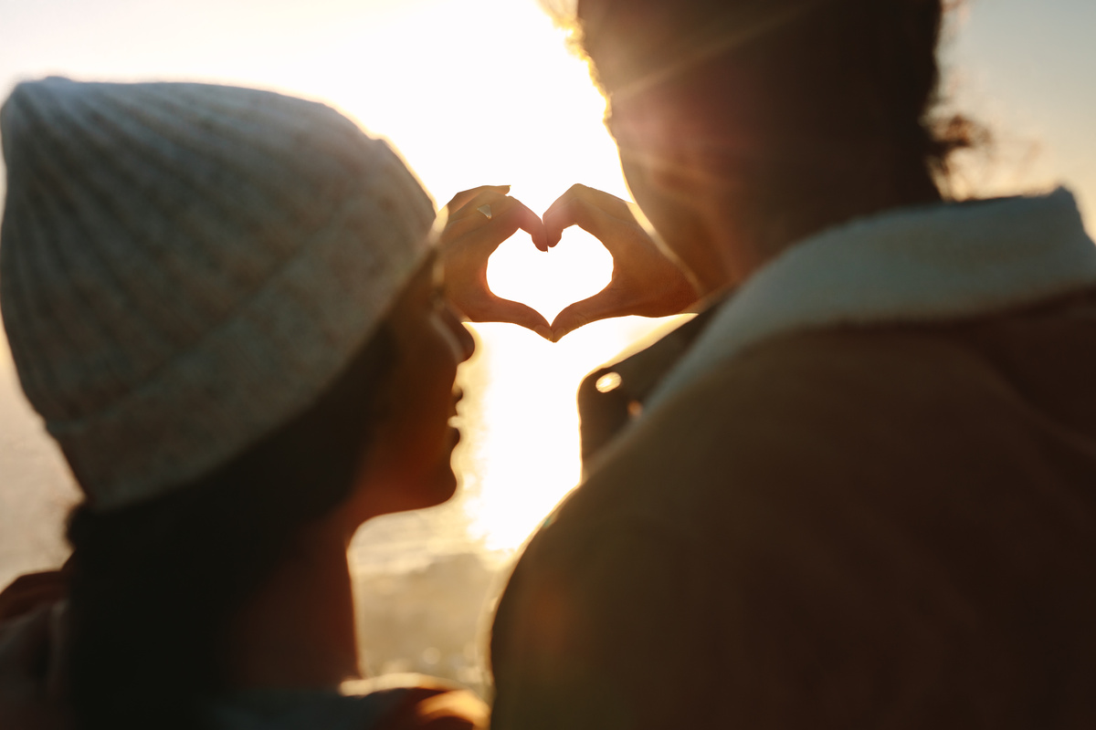 Homem e mulher juntando suas mãos em um coração em frente à luz do sol.