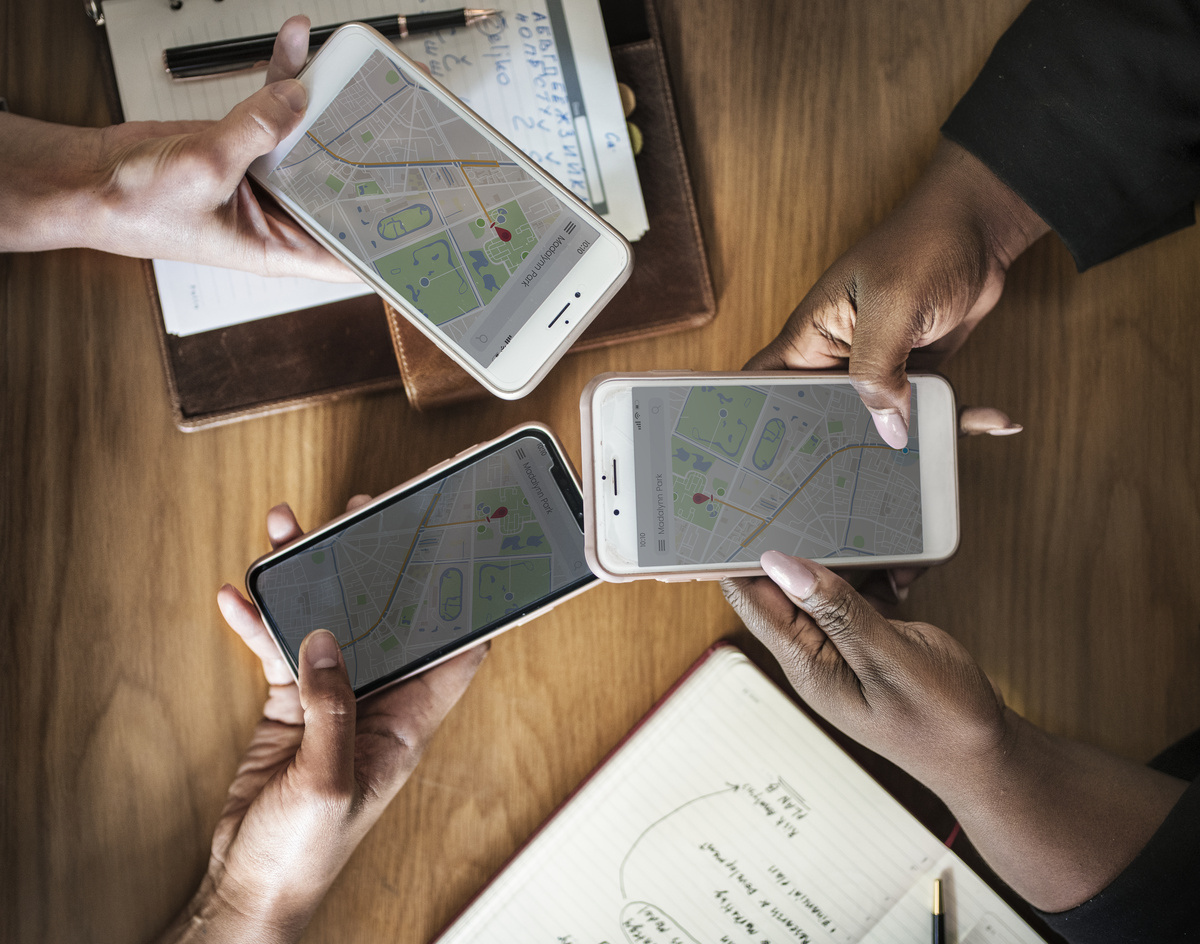 Três mãos segurando celulares que mostram GPS na tela, parecendo procurar por um endereço.
