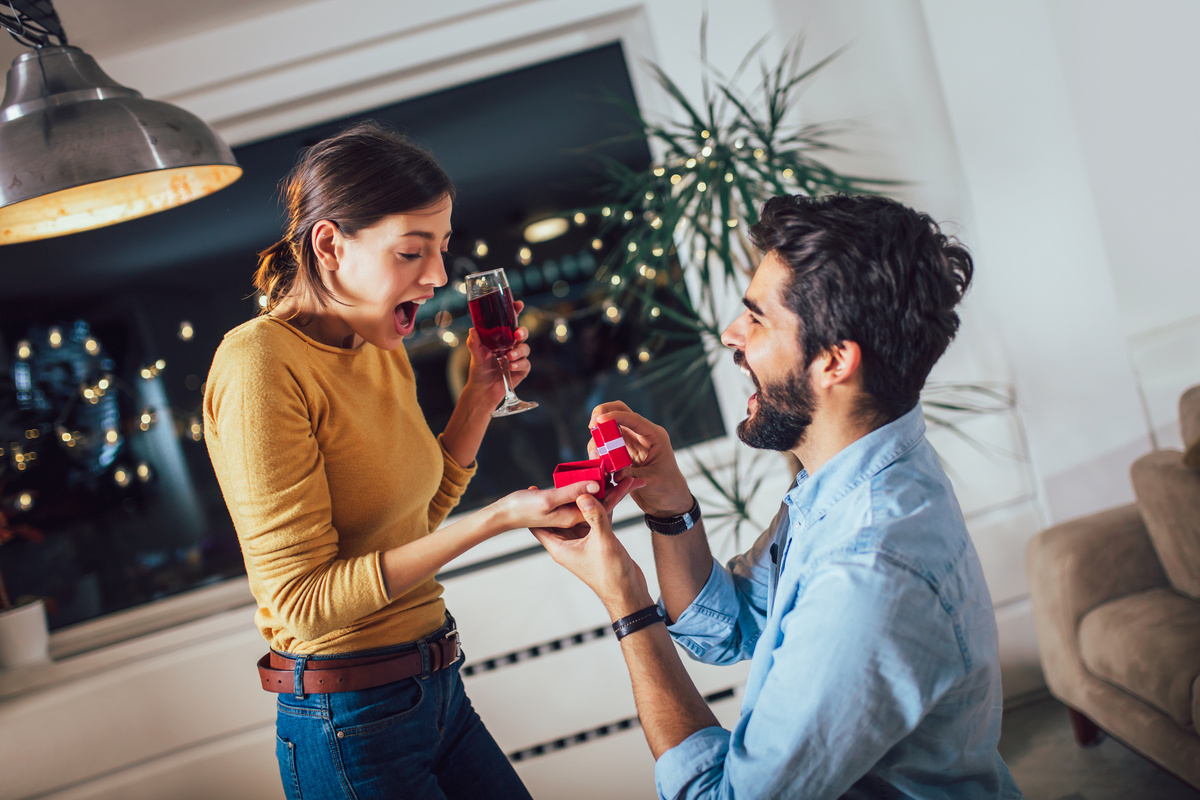 Mulher em pé, sorrindo e com taça de vinho na mão, enquanto homem se encontra ajoelhado de frente para ela, presenteando-a com uma joia. 