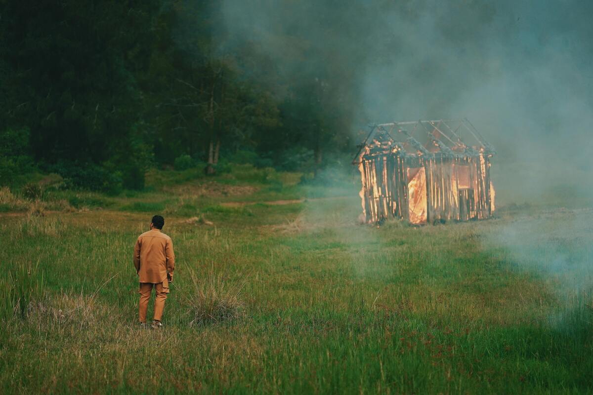 Homem em pé em um campo gramado observando uma casa de palha pegar fogo.