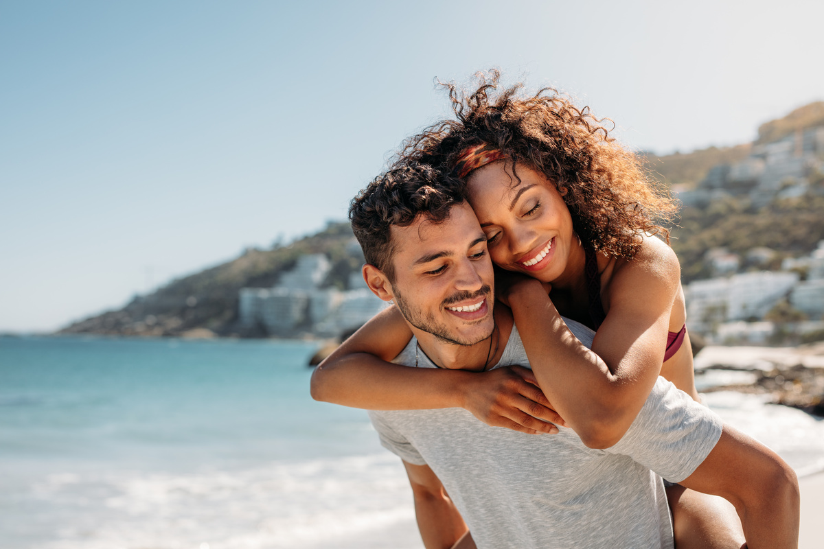 Em beira da praia durante dia ensolarado, homem carregando mulher nas costas enquanto ambos sorriem, representando a combinação do casal Gêmeos e Aquário.