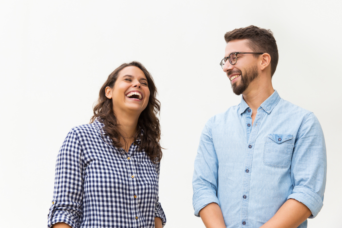 Homem e mulher rindo, um ao lado do outro, em local de fundo branco, representando Aquário e Gêmeos com base no gênero.