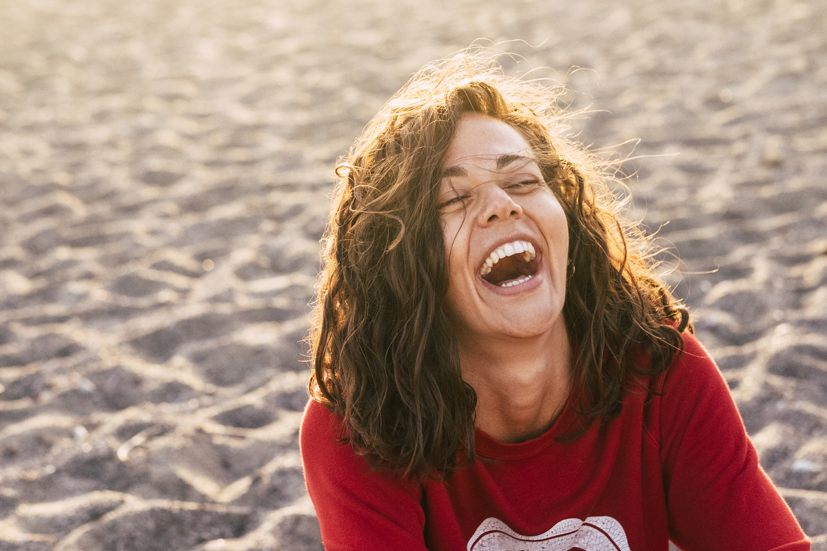 Mulher sentada na areia enquanto gargalha, representando sonhar com rir. 