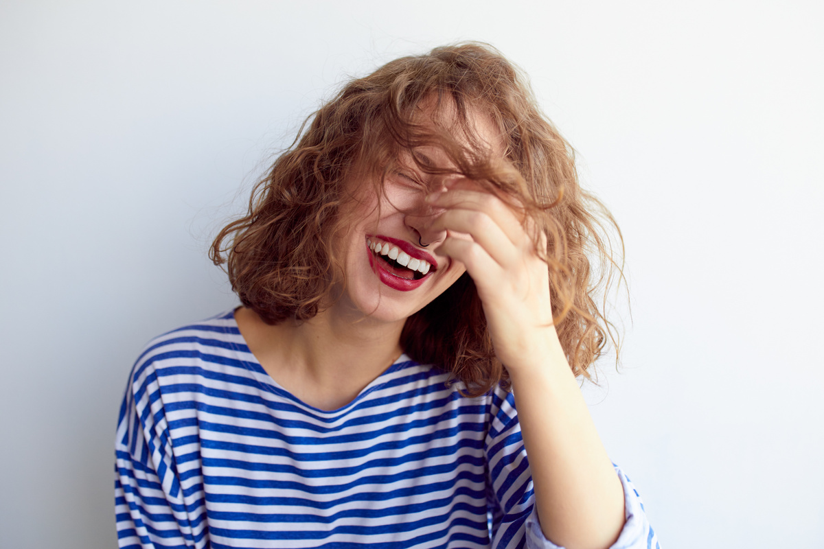 Mulher tirando os cabelos ondulados de frente do rosto enquanto ri, representando sonhar com rir.