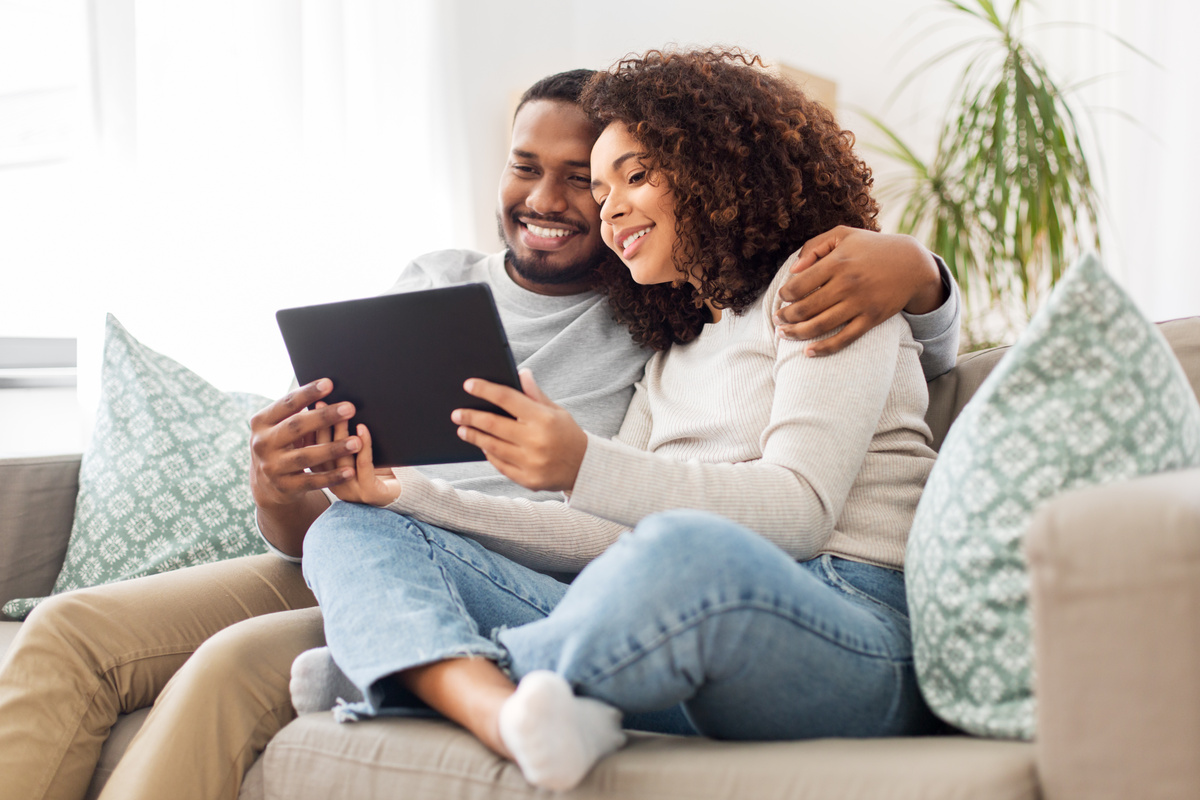 Homem e mulher sorridentes sentados em sofá olhando para tela de tablet.