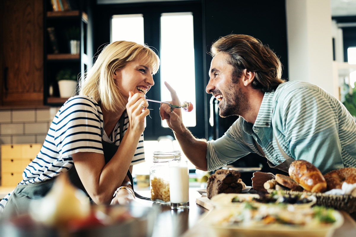Homem tocando nariz de mulher enquanto ela segura o garfo para dar comida a ele e ambos sorriem, representando a combinação Aquário e Câncer.