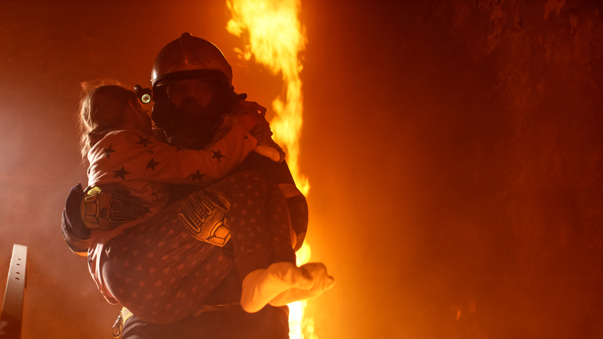 Bombeiro carregando criança em colo, com cenário atrás dele em chamas, representando o ato de salvar alguém.