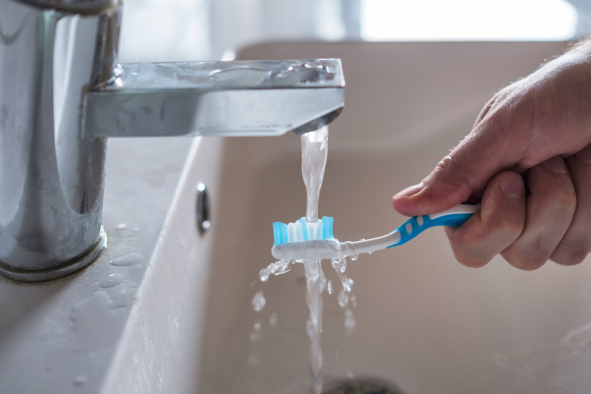 Mão segurando escova de dentes embaixo de água corrente da torneira. 
