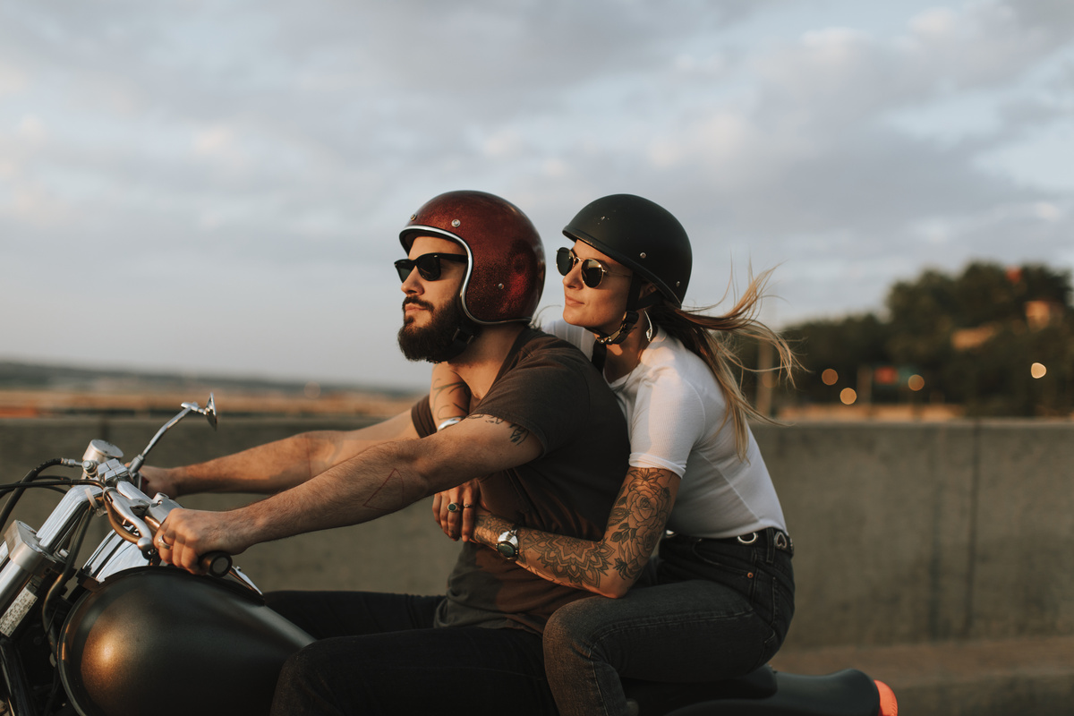 Homem dirigindo moto enquanto mulher, sentada na garupa, o abraça por trás.