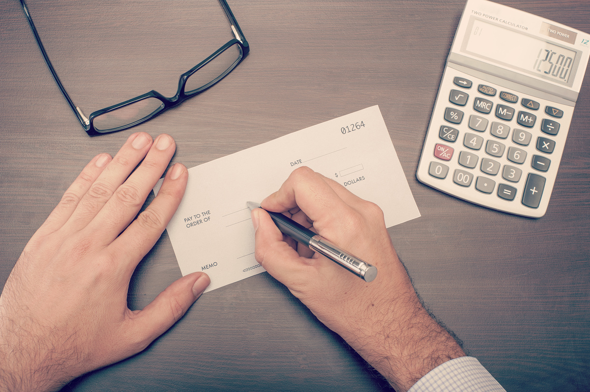 Homem preenchendo cheque com calculadora e óculos ao lado