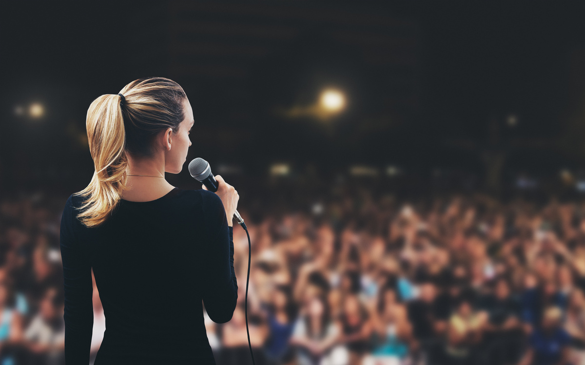 Imagem de costas de mulher loira, que segura um microfone e apresenta sua palestra em um auditório escuro e cheio de pessoas.