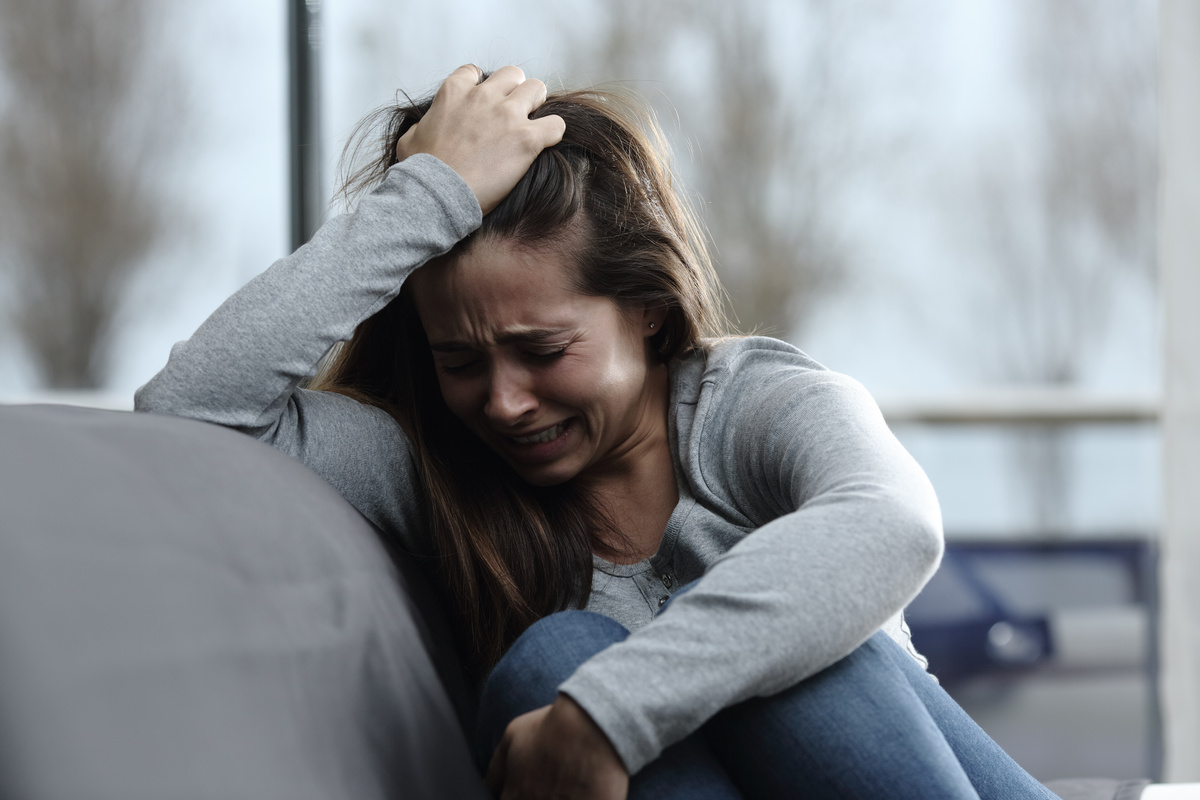 Mulher sentada em sofá enquanto chora e tem a mão na cabeça, representando a vítima da amarração amorosa sofrendo os seus efeitos.